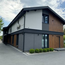 ELTAR Konrad Lamprecht - Nowoczesny, dwukondygnacyjny dom z elewacją w odcieniach szarości, bieli i drewna. Widoczne elementy wykończeniowe i zadbany teren wokół budynku.