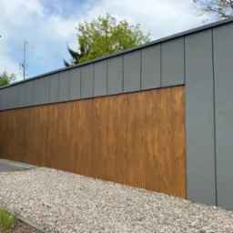 Nowoczesna elewacja budynku z drewnianymi panelami i szarą, gładką powierzchnią. Uwagę przyciąga kontrast materiałów i staranne wykończenie detali.