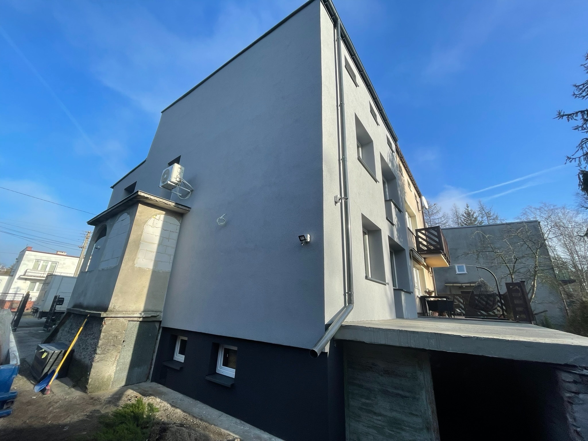 Odnowiona elewacja budynku wielorodzinnego w odcieniach szarości, z widocznym fragmentem balkonu i klimatyzatorem na ścianie, uchwycona w słoneczny dzień z perspektywy żabiej