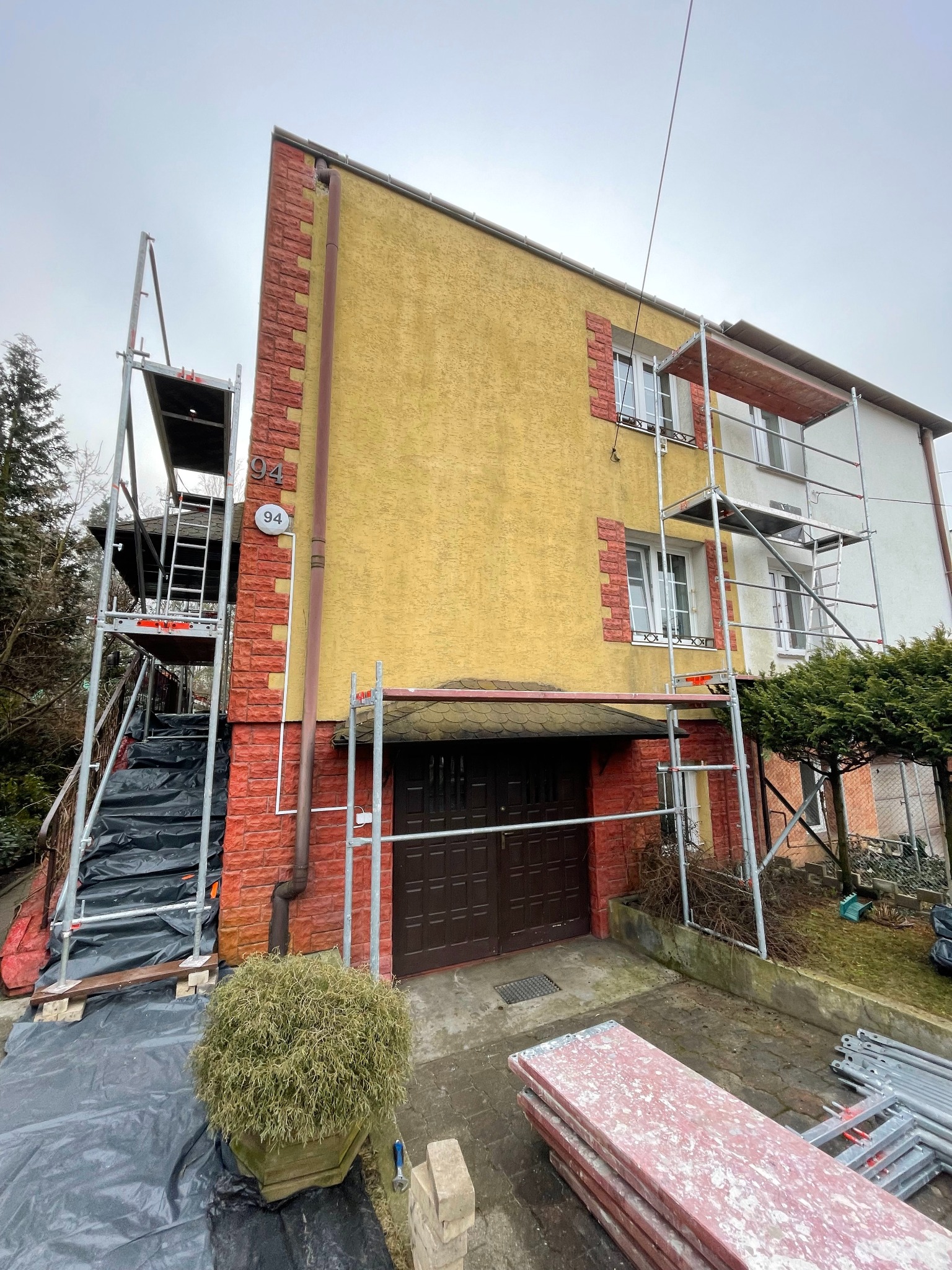 Dwupiętrowy dom z żółtą elewacją i czerwoną cegłą, otoczony rusztowaniami podczas prac remontowych, widoczny numer domu 94.