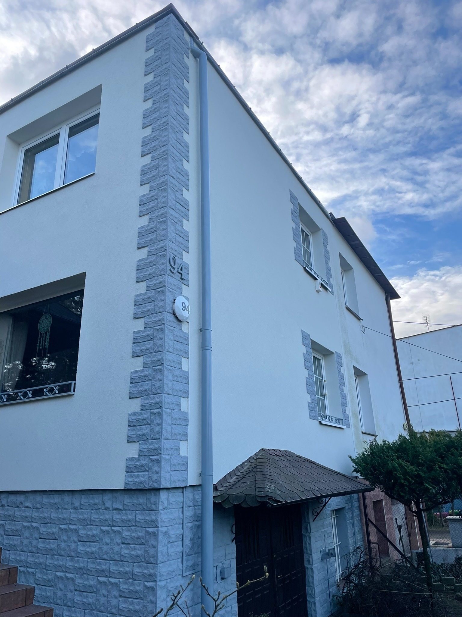 Dwupiętrowy dom z białą elewacją, ozdobiony szarymi panelami imitującymi kamień wokół okien i na narożnikach, z numerem domu '94' na elewacji.