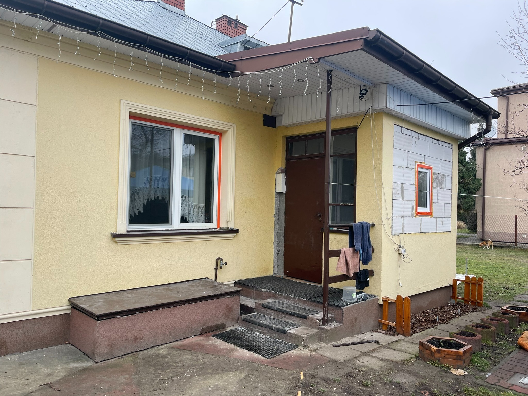 Żółta elewacja domu jednorodzinnego z białymi oknami i brązowymi drzwiami wejściowymi, widoczne fragmenty ocieplenia z białych bloczków, dekoracyjne lampki na dachu, teren wokół domu częściowo...