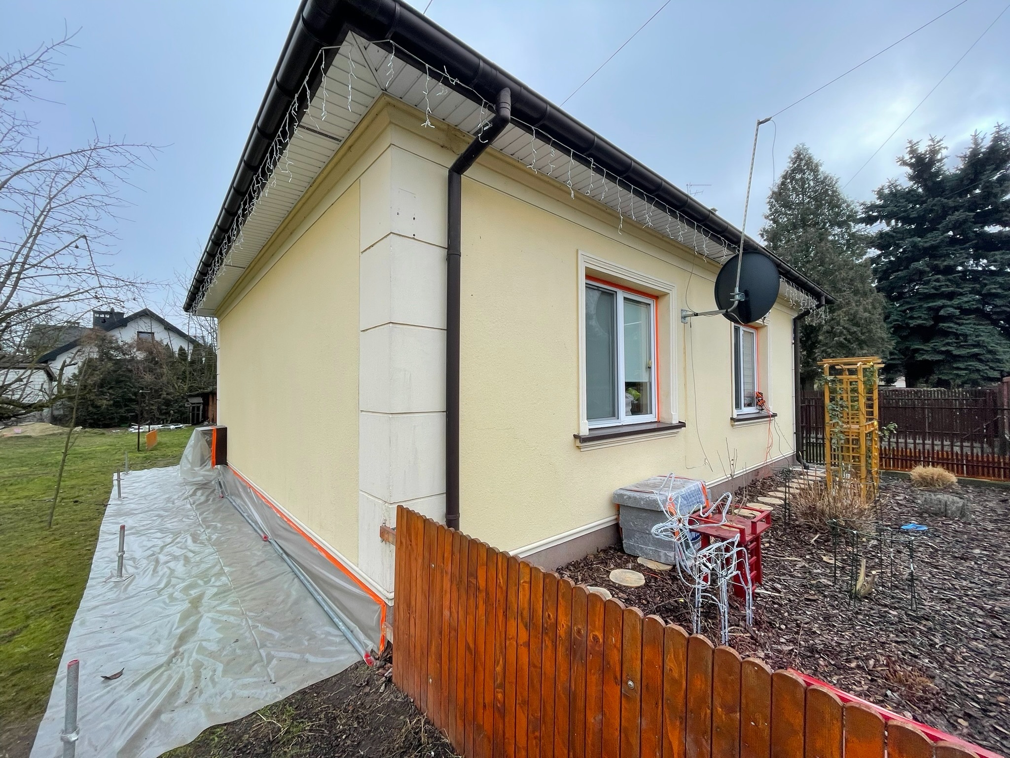Żółta elewacja domu jednorodzinnego po remoncie, z dekoracyjnymi elementami architektonicznymi, zabezpieczona folią ochronną u podstawy, widoczna antena satelitarna i drewniany płot.