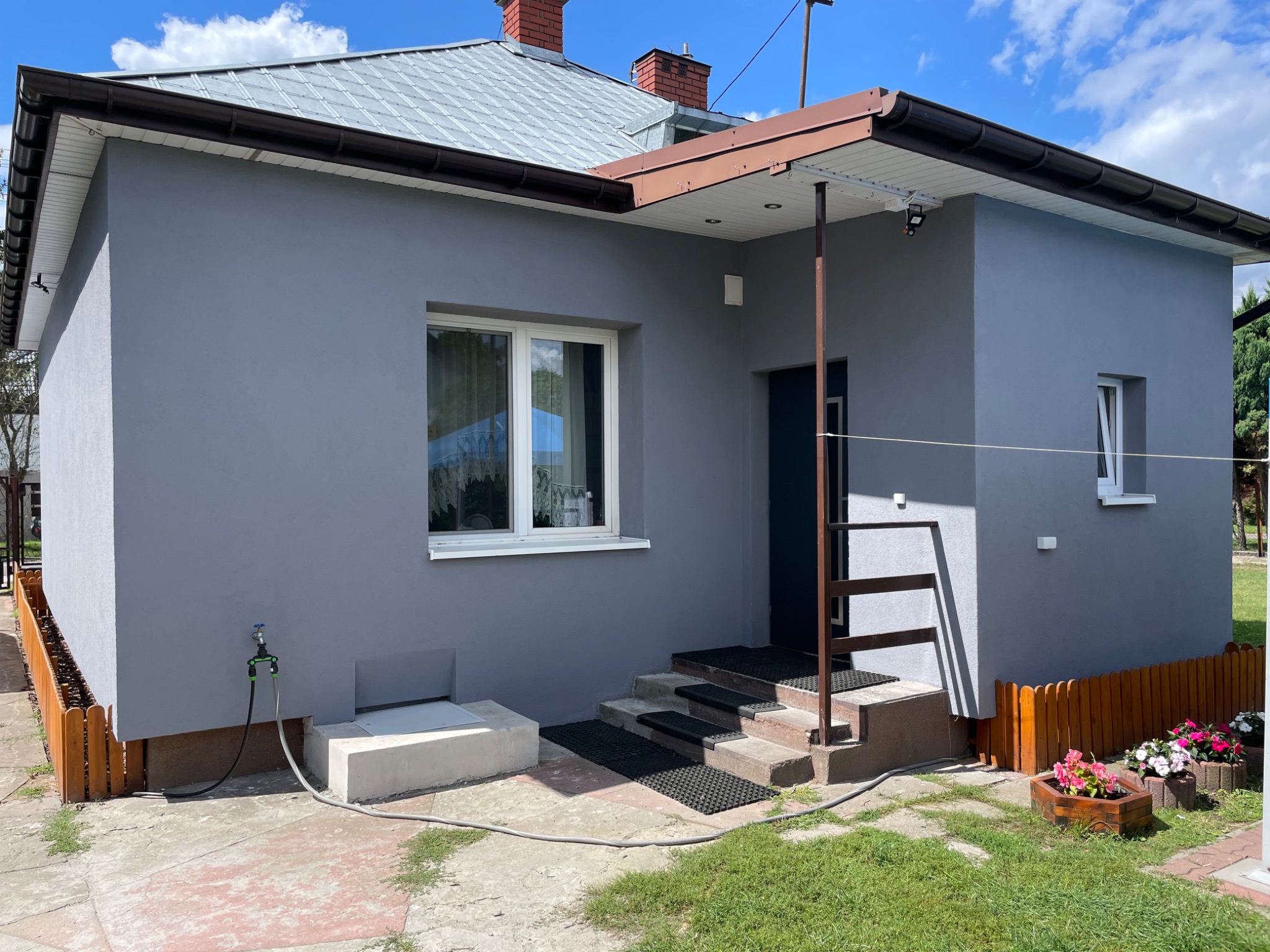 Szary dom jednorodzinny z brązowymi rynnami i schodami wejściowymi. Świeżo pomalowana elewacja w słoneczny dzień, widoczne kwiaty w donicach i fragment ogrodzenia.