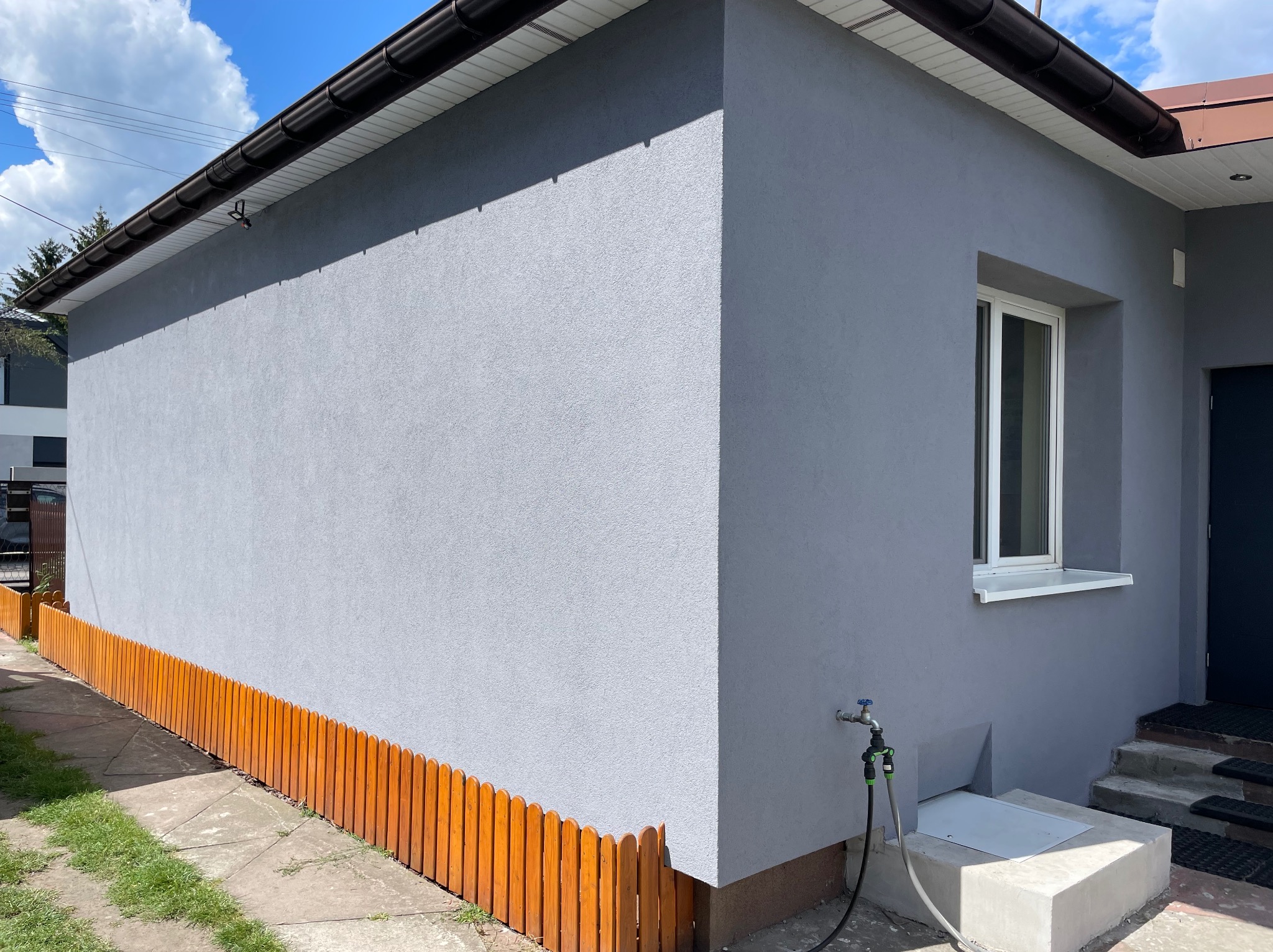 Szara elewacja budynku z oknem i brązowym ogrodzeniem. Widoczny fragment dachu z rynnami, trawnik i kran z wężem.