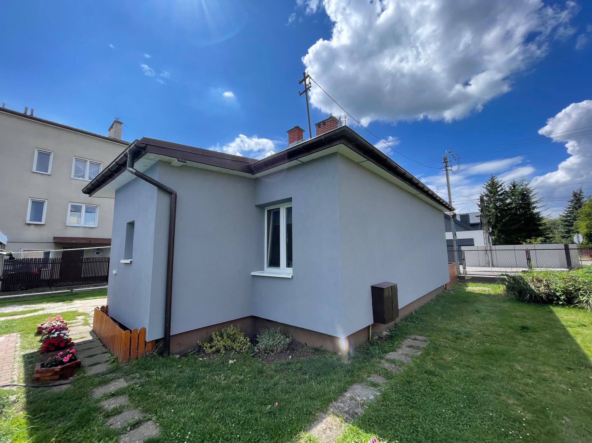 Szary dom jednorodzinny po renowacji elewacji, widok z boku na tle błękitnego nieba z chmurami. Widoczne detale: brązowe rynny, białe okna, drewniane ogrodzenie.