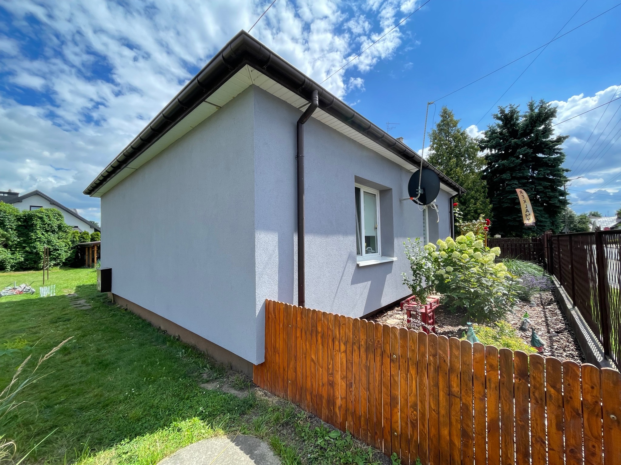Jasnoszary dom jednorodzinny po remoncie elewacji, z brązowymi rynnami i drewnianym płotem. Widok z boku na tle błękitnego nieba z chmurami.