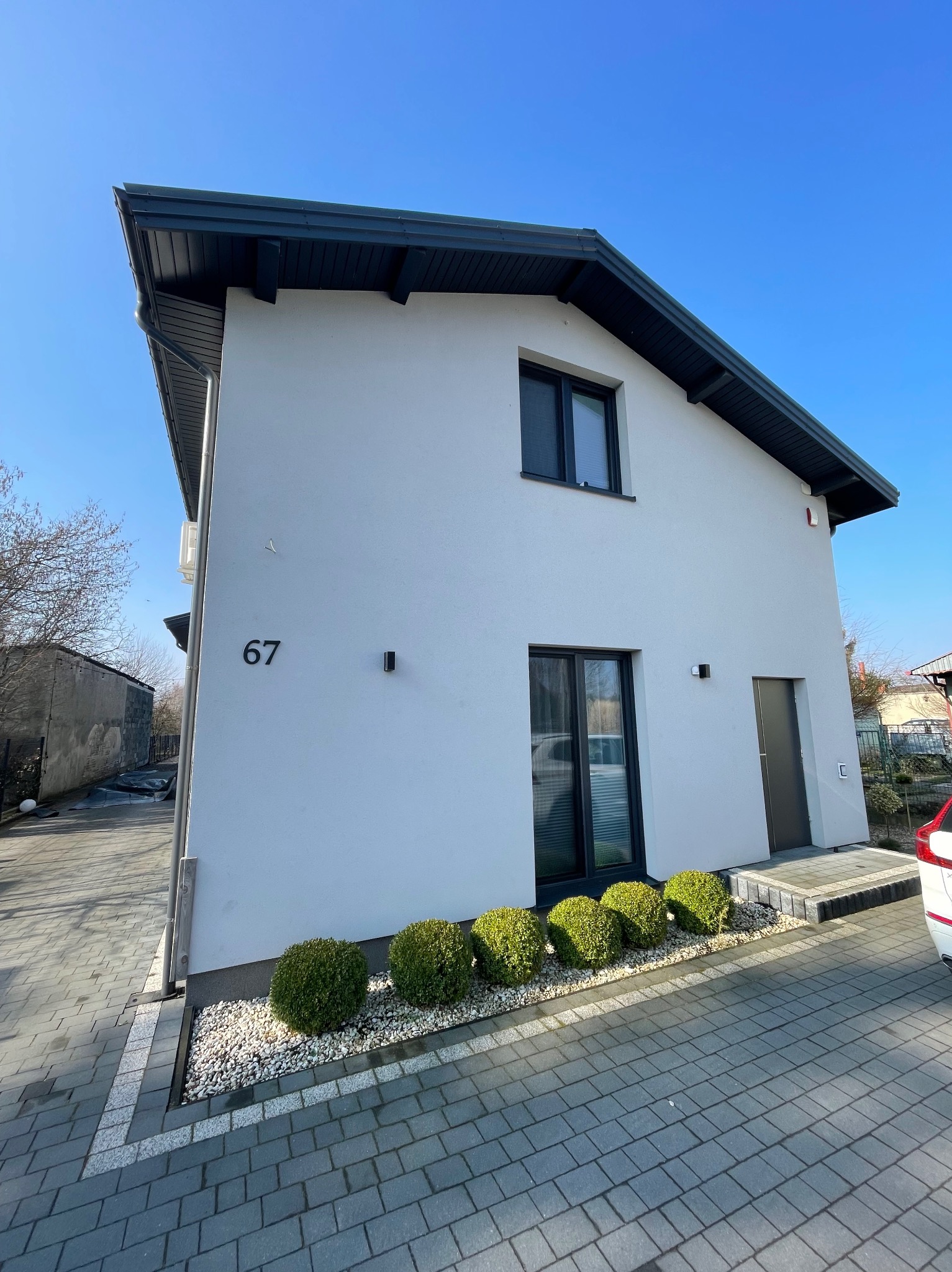 Biały, dwukondygnacyjny dom z numerem 67, czarną stolarką i dachem, ocieplony, z równo przyciętymi krzewami i szarym brukiem na podjeździe, w słoneczny dzień.