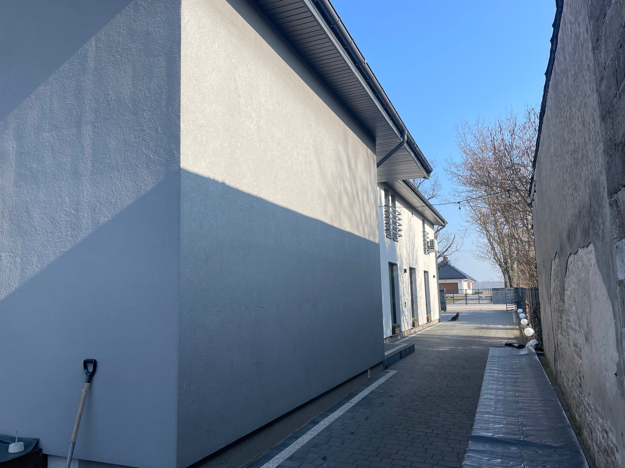 Świeżo otynkowana, szara ściana budynku z widoczną fakturą, z łopatą opartą o ścianę. W tle widoczny fragment chodnika z kostki i niebieskie niebo.
