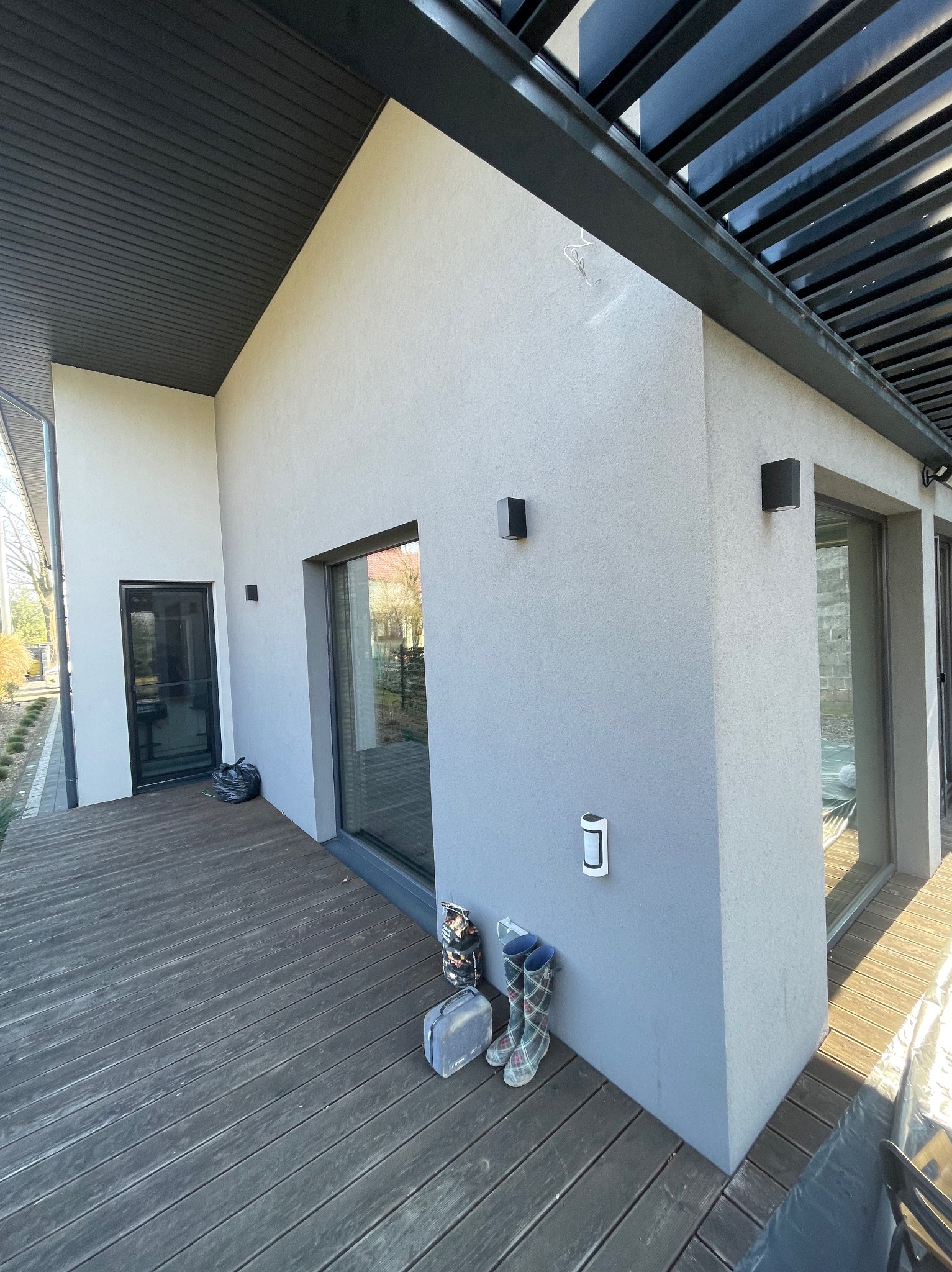 Jasnoszara elewacja domu z drewnianym tarasem i czarnymi elementami wykończeniowymi. Widoczne okna, oświetlenie zewnętrzne i buty robocze przy ścianie.