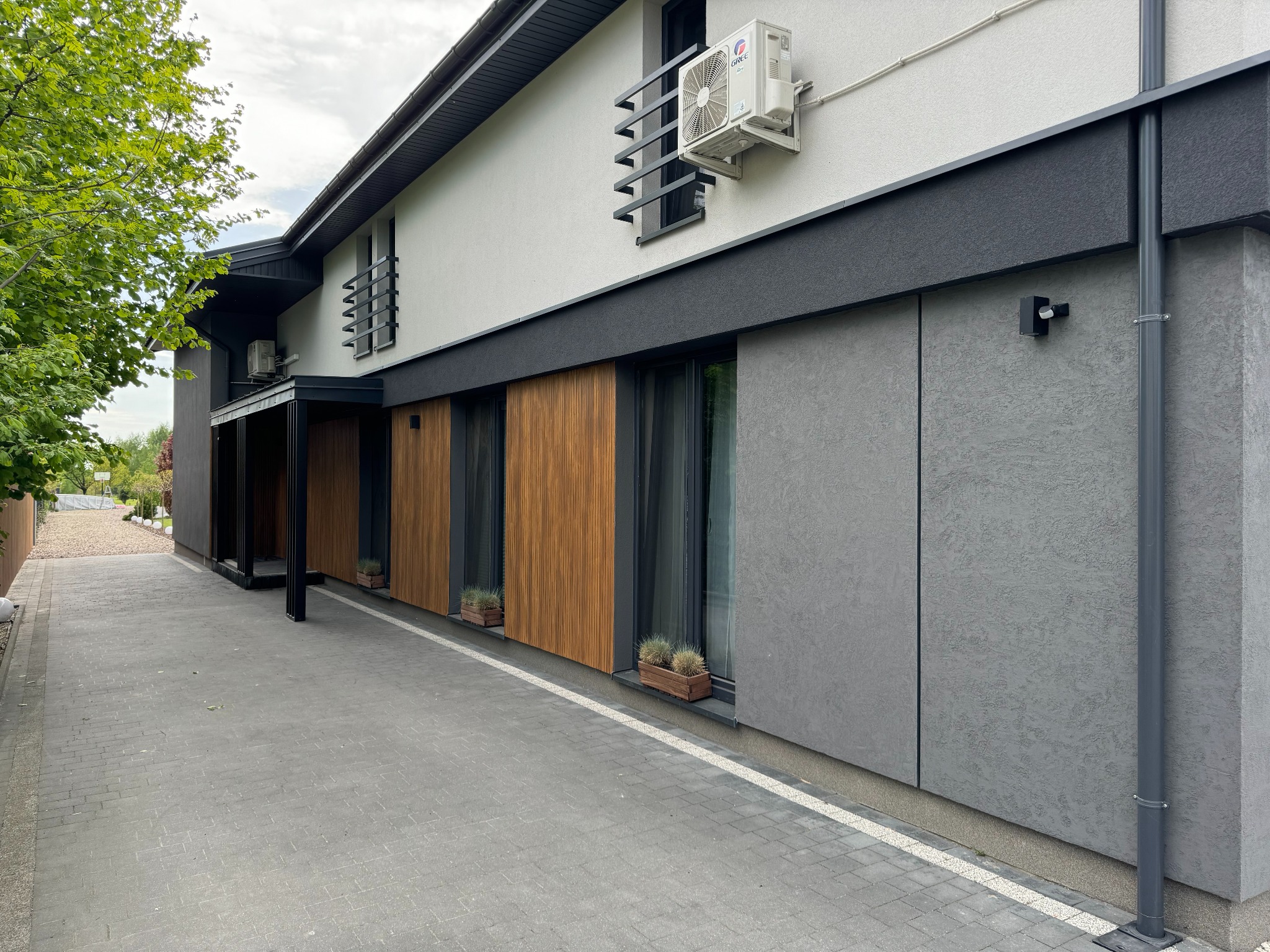 Nowoczesna elewacja domu z elementami drewna, szarego tynku i białej ściany. Widoczny klimatyzator, ozdobne barierki okienne i kostka brukowa na podjeździe.