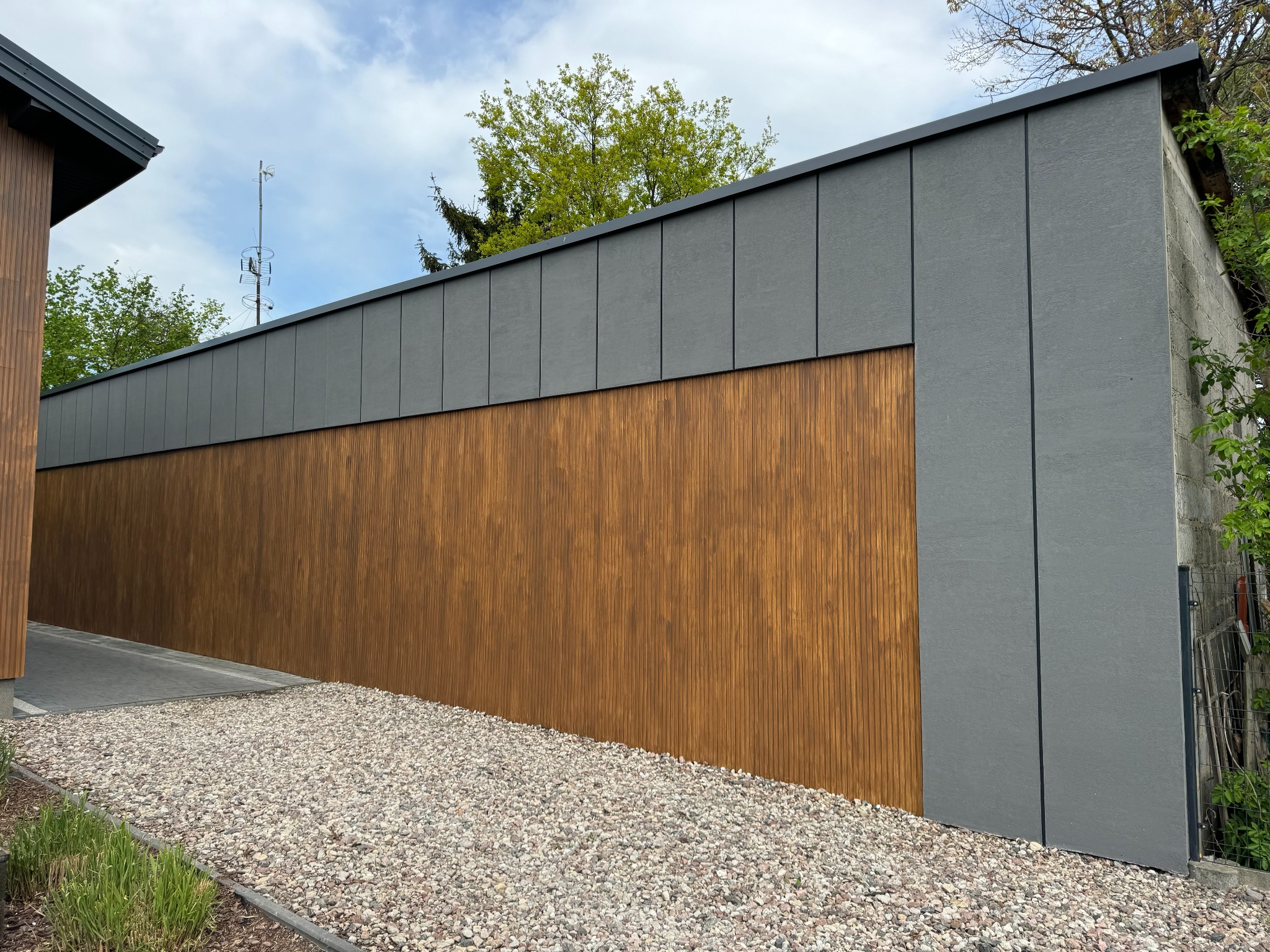 Nowoczesna elewacja budynku z drewnianymi panelami i szarą, gładką powierzchnią. Uwagę przyciąga kontrast materiałów i staranne wykończenie detali.