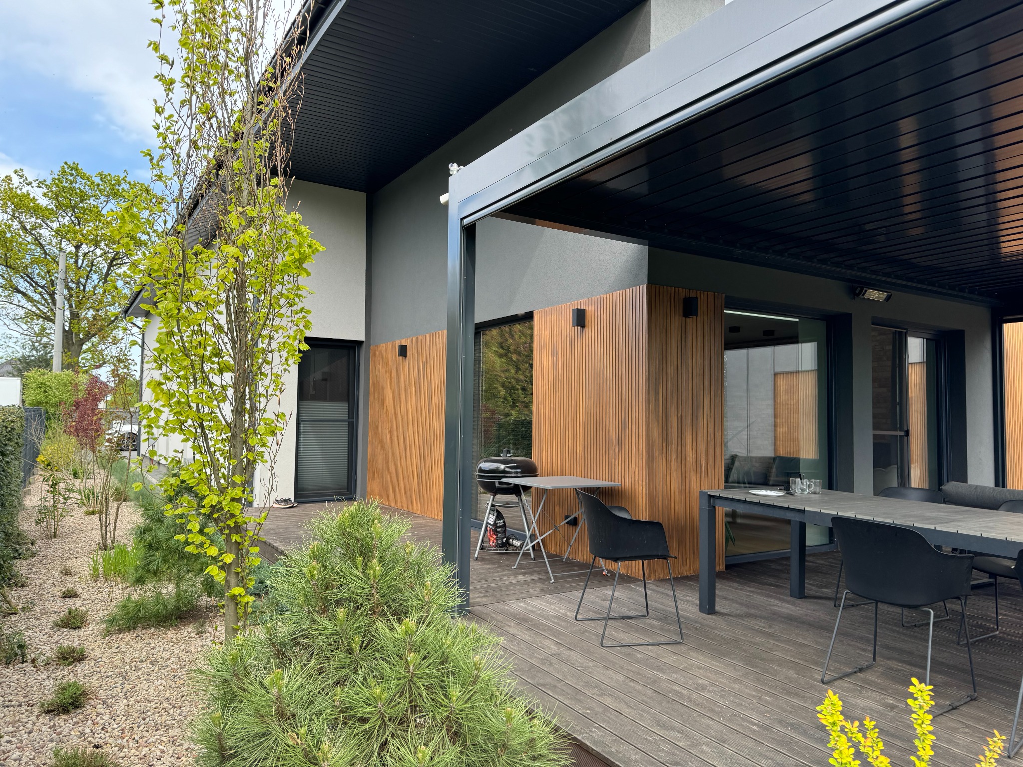 Nowoczesny dom z drewnianą elewacją i tarasem. Widoczne meble ogrodowe, grill. W tle zieleń ogrodu i błękitne niebo. Całość w stonowanej kolorystyce.