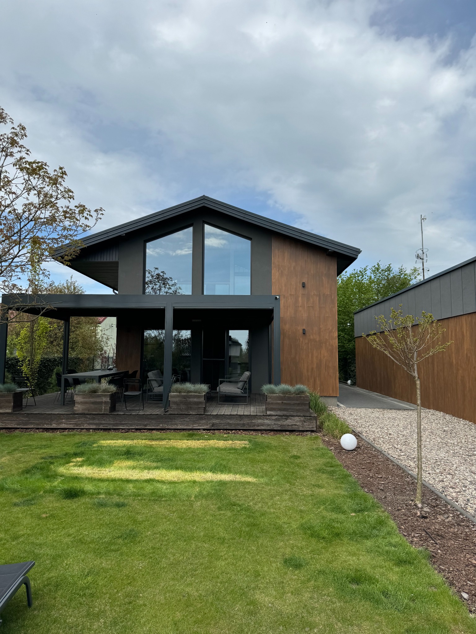 Nowoczesny dom jednorodzinny z drewnianą elewacją i dużymi przeszkleniami, otoczony zadbanym trawnikiem i elementami małej architektury.