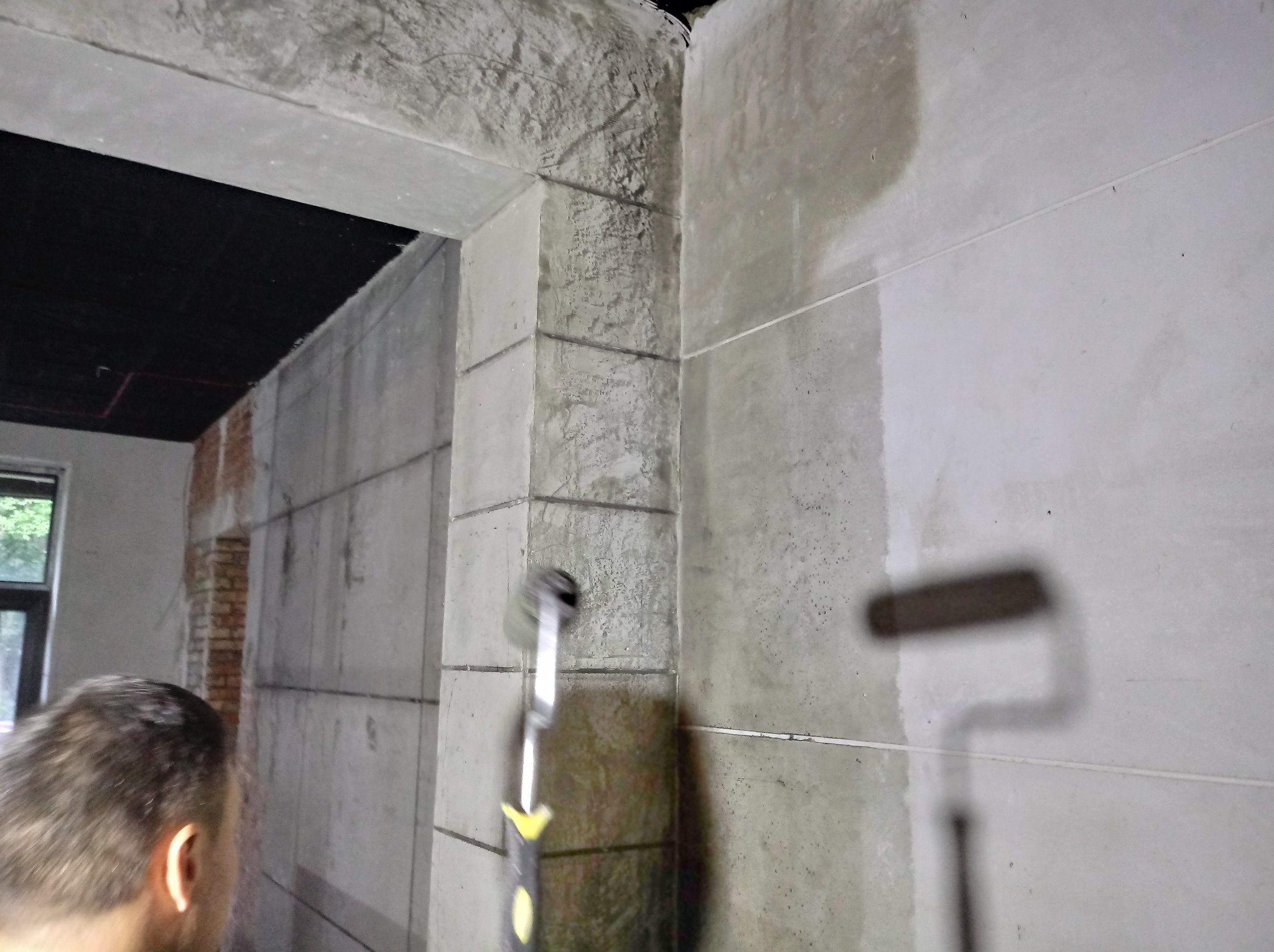 Malowanie szarej ściany wałkiem z żółtymi akcentami na uchwycie, widoczny fragment osoby malującej i surowy stan wykończenia wnętrza z oknem w tle.