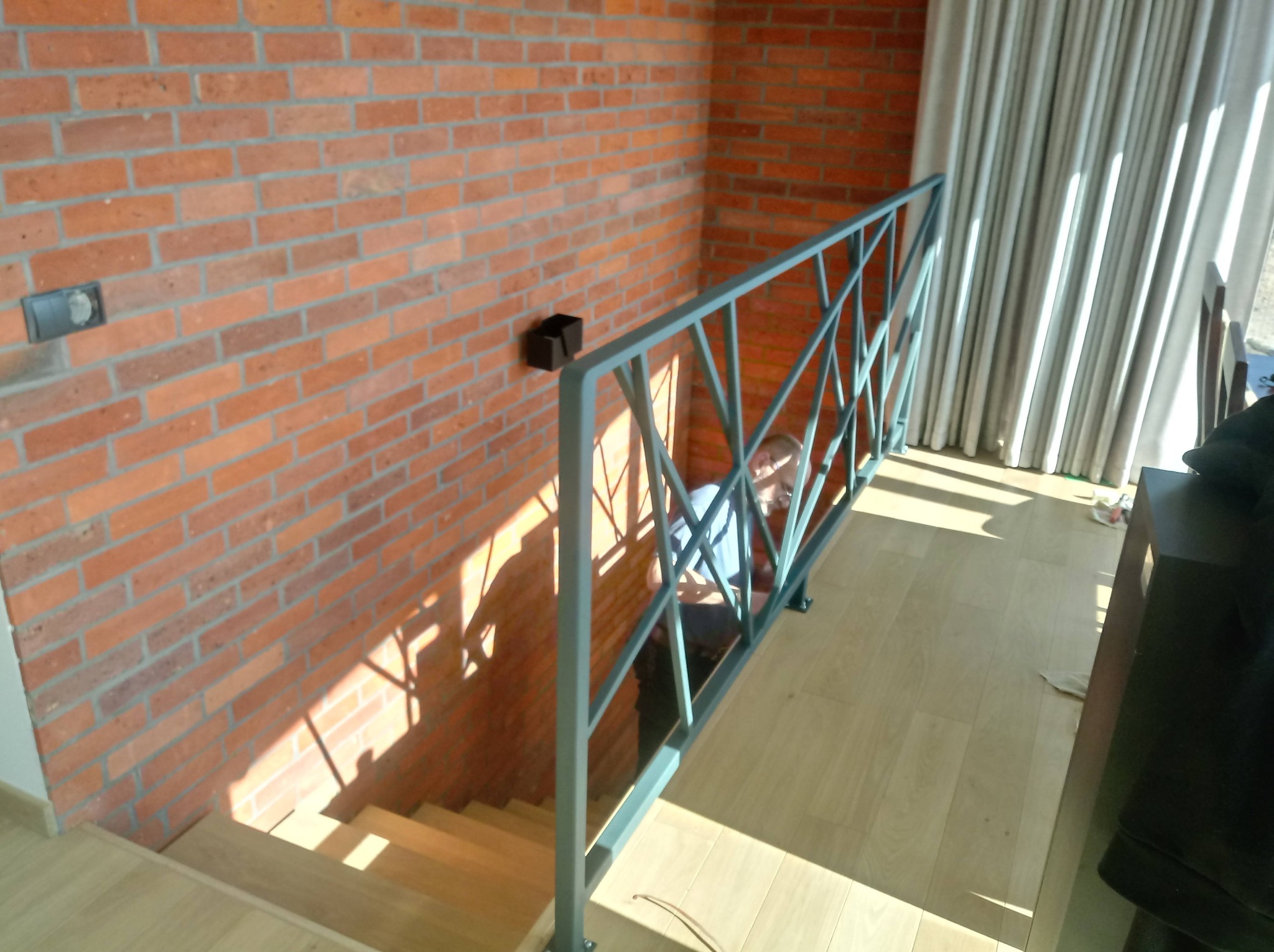 Schody z drewnianymi stopniami i szarą metalową balustradą o geometrycznym wzorze, ściana z czerwonej cegły z widocznymi przewodami elektrycznymi, jasne drewniane panele podłogowe i wpadające...