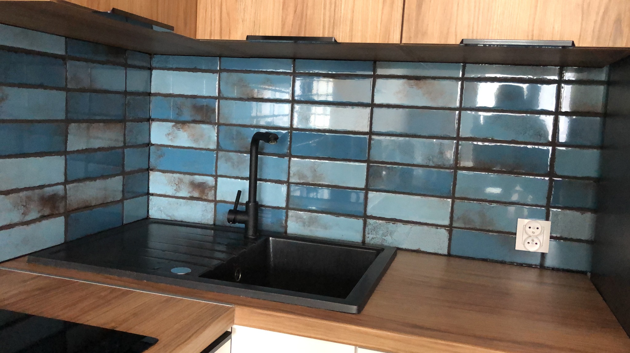 Błękitne, prostokątne płytki z efektem rdzy na ścianie nad czarnym, granitowym zlewem i czarną baterią w kuchni z drewnianym blatem.