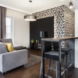 Nowoczesny salon z szarą sofą, geometryczną tapetą i drewnianym blatem baru z hokerami. Minimalistyczne oświetlenie i ciemna podłoga dodają elegancji.