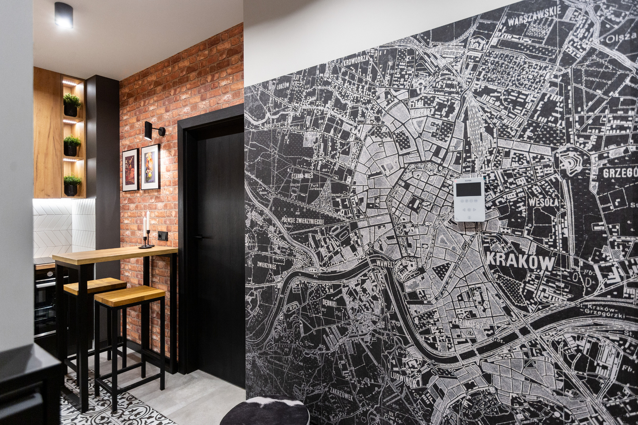 Nowoczesna kuchnia z ceglaną ścianą, wysokim stołem barowym i krzesłami, na ścianie tapeta z mapą Krakowa, oświetlenie punktowe i dekoracyjne rośliny w podświetlanej wnęce.