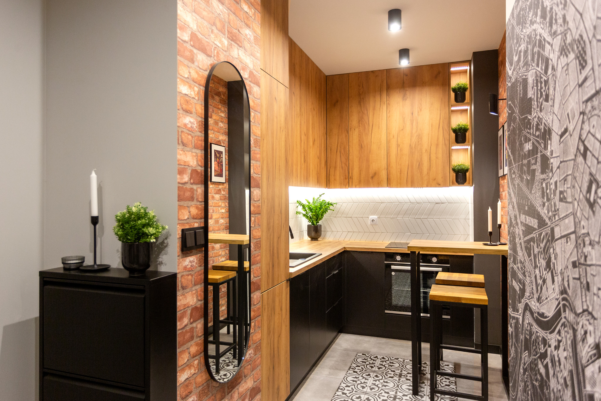 Nowoczesna kuchnia z drewnianymi frontami szafek i czarnymi elementami, ceglana ściana z owalnym lustrem, ściana z tapetą w mapę miasta, wysokie stołki barowe przy blacie, wzorzysta podłoga.