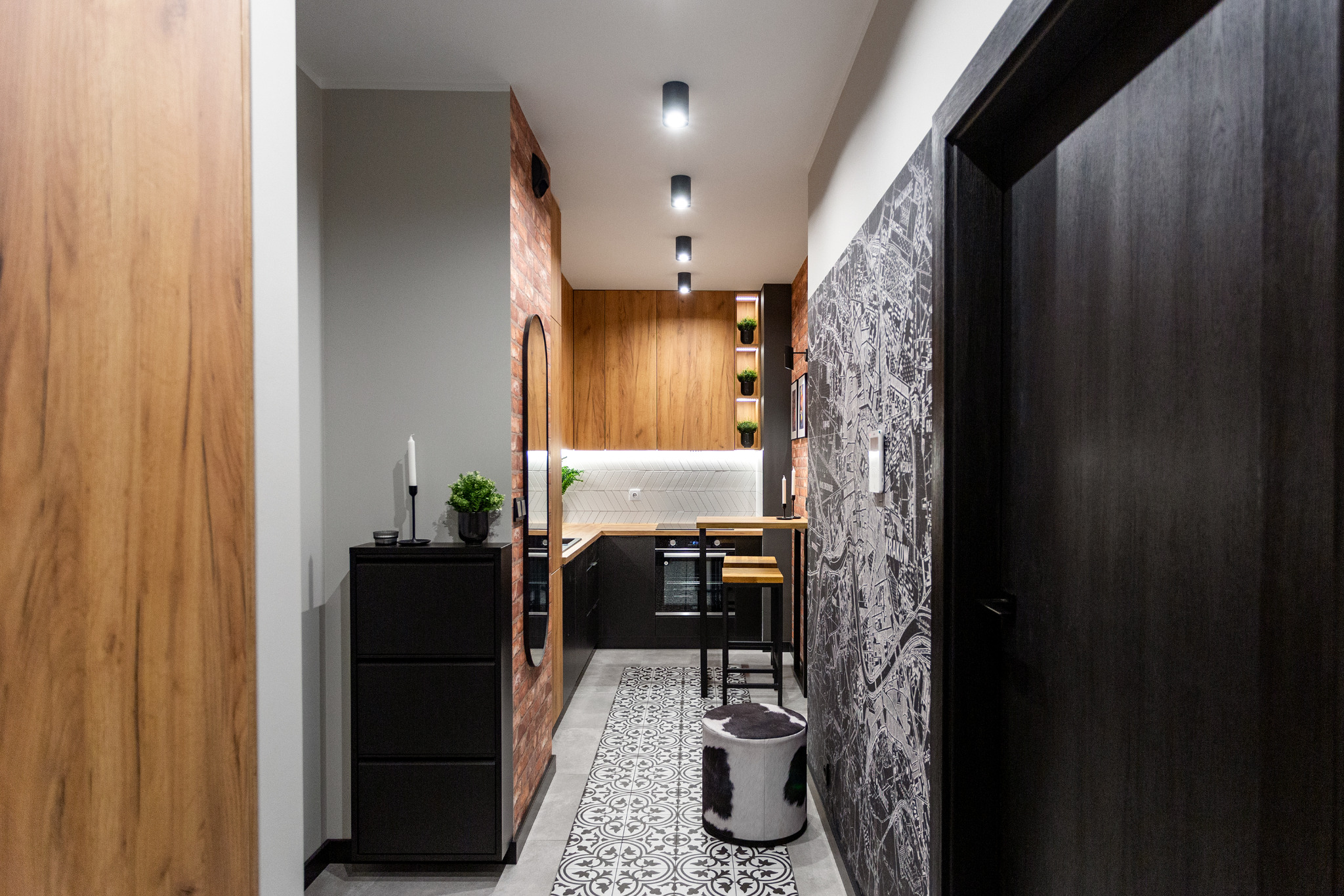 Wąski korytarz z aneksem kuchennym, ceglana ściana, tapeta z mapą miasta, czarne meble, wzorzysta podłoga, oświetlenie punktowe.