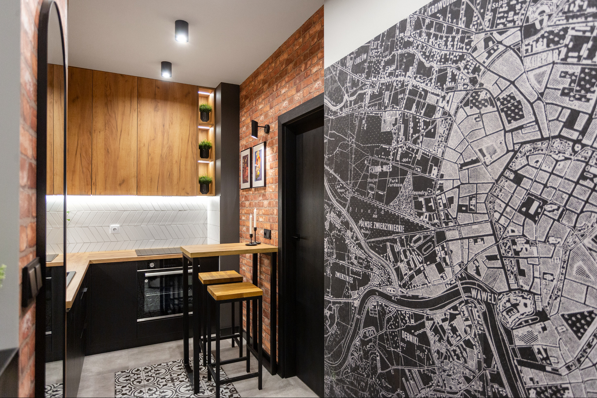 Nowoczesna kuchnia z czarnymi szafkami, drewnianym blatem i ścianą z cegły, uzupełniona wysokim stołem barowym i krzesłami, a także tapetą z mapą Krakowa.