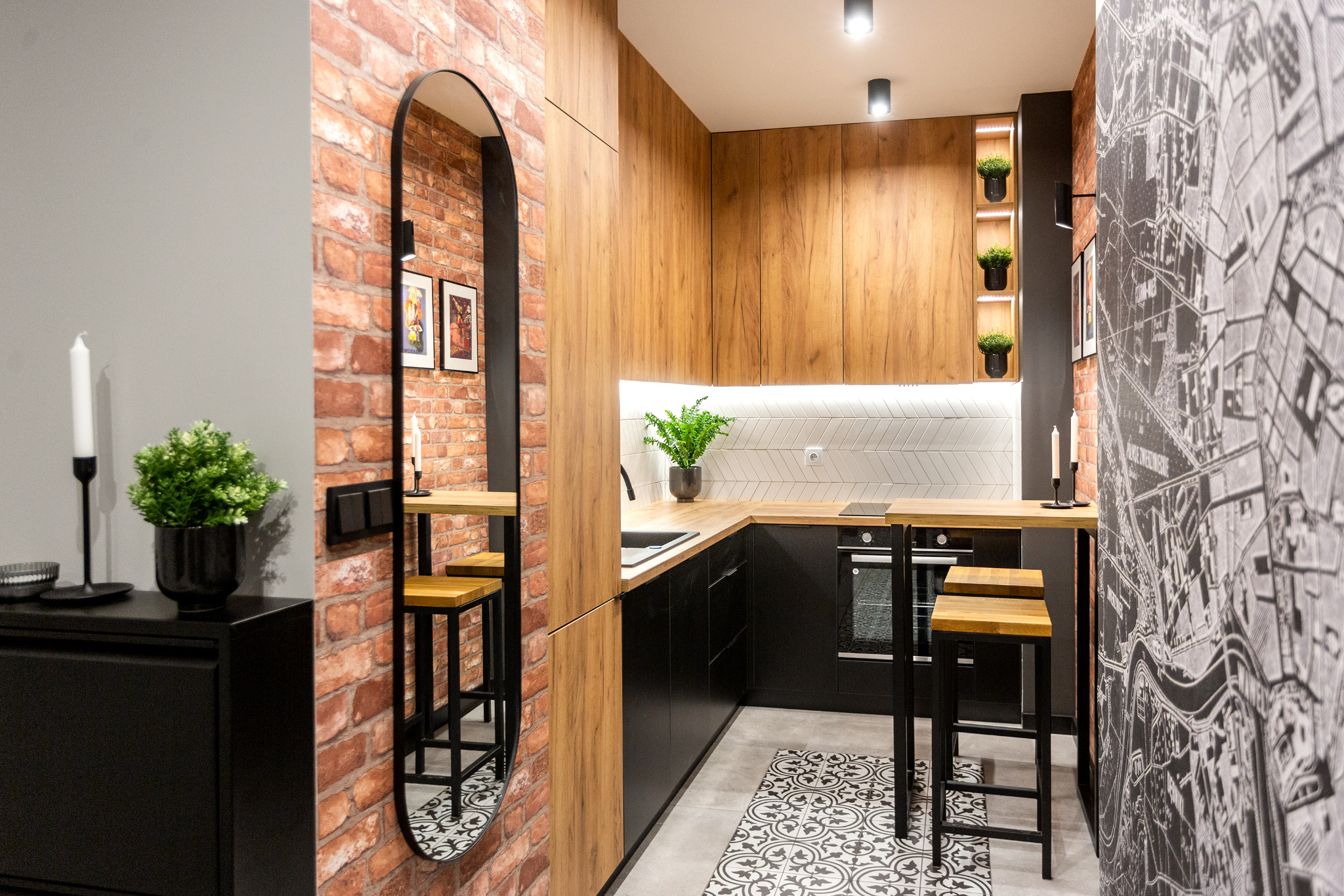 Nowoczesna kuchnia z drewnianymi szafkami, czarnymi frontami i ceglaną ścianą z owalnym lustrem, uzupełniona geometryczną podłogą i tapetą z mapą miasta.