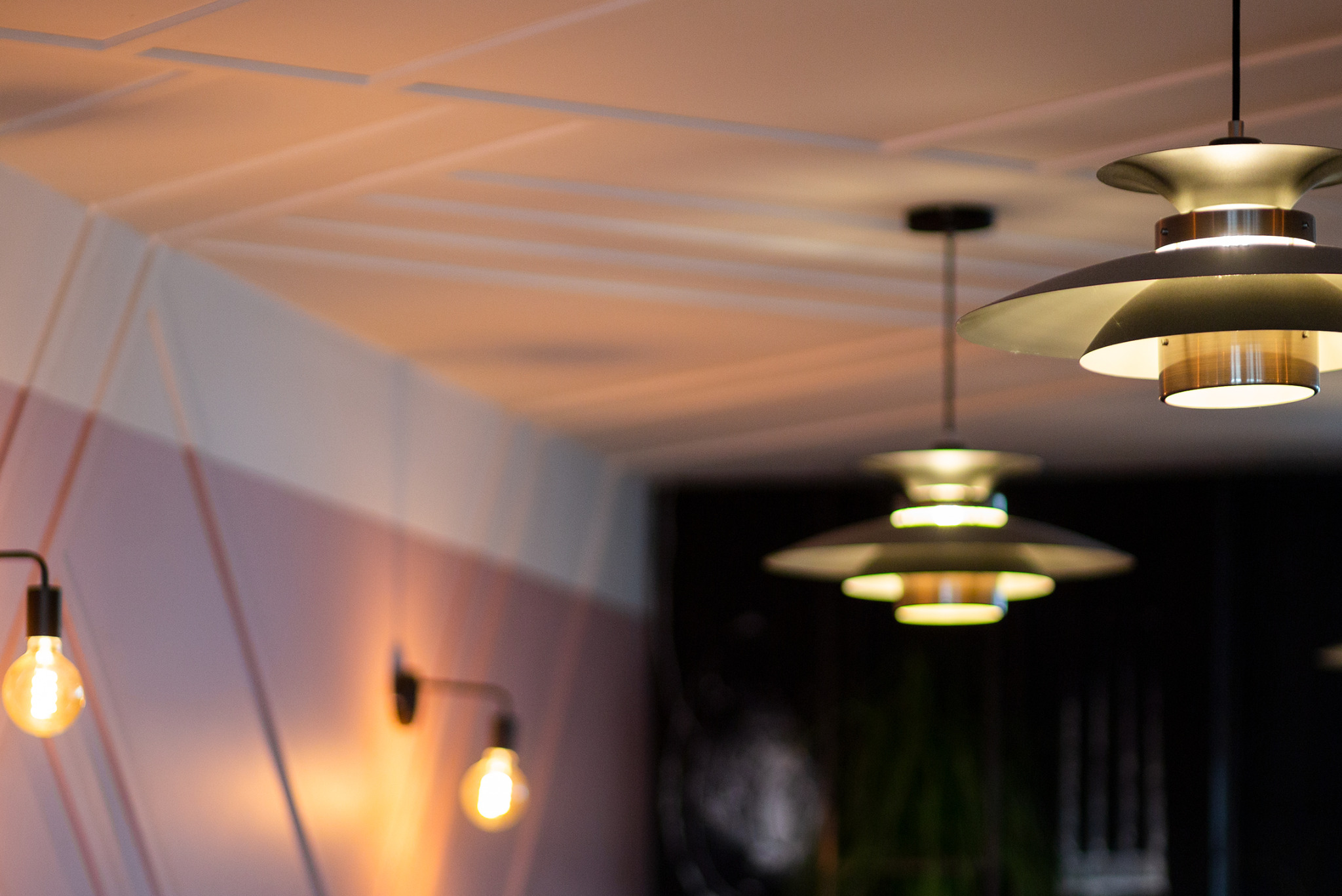 Nowoczesne oświetlenie wnętrza z trzema lampami wiszącymi o warstwowej konstrukcji i ciepłym świetle żarówek Edisona na ścianie z geometrycznym wzorem.