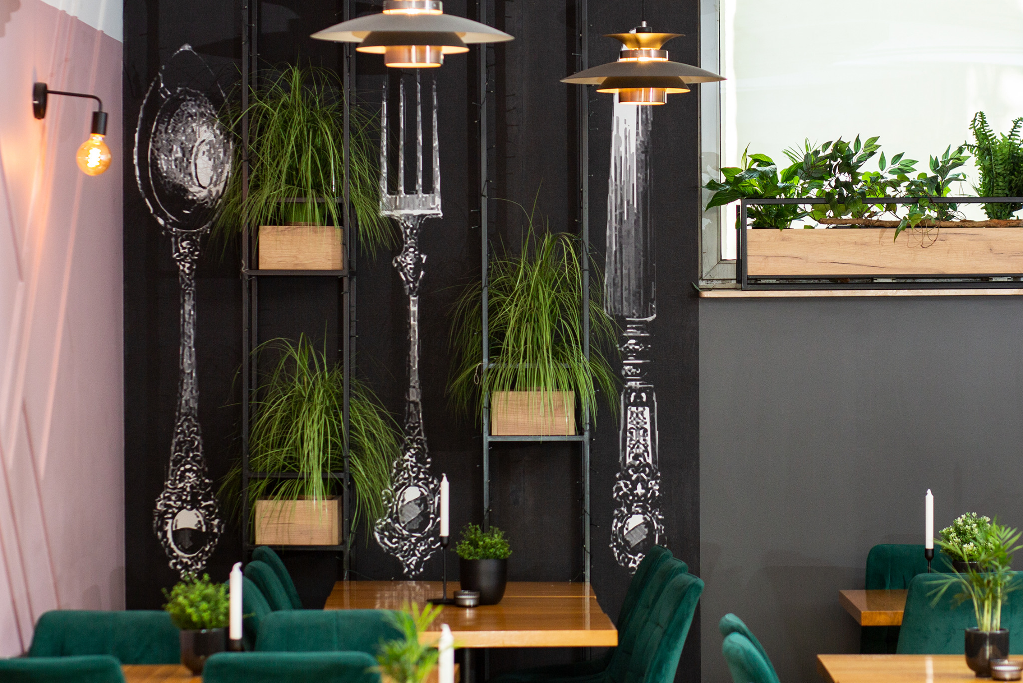 Wnętrze restauracji z graficznymi motywami sztućców na ścianie, roślinami w drewnianych skrzynkach na metalowych półkach, zielonymi krzesłami welurowymi i wiszącymi lampami w stylu retro.