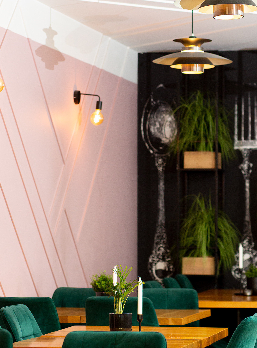 Wnętrze restauracji z elementami loftowymi: pastelowa ściana z geometrycznym wzorem, czarna ściana z grafiką sztućców, drewniane stoły i zielone krzesła z weluru, oświetlenie punktowe i wiszące lampy.