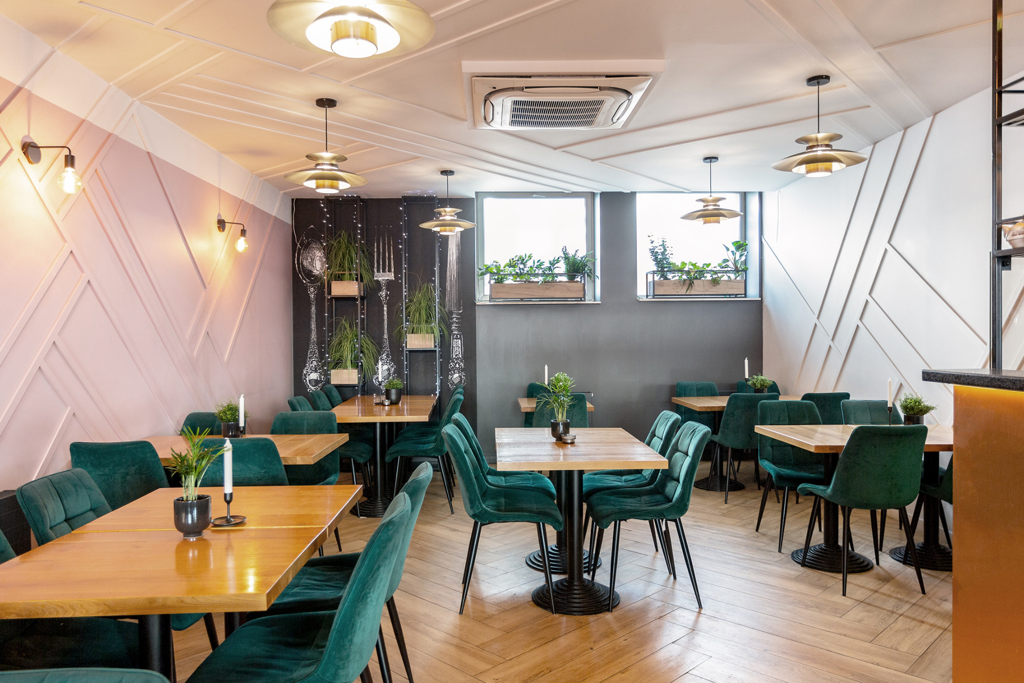 Jasne wnętrze restauracji z drewnianymi stolikami, zielonymi krzesłami z weluru i dekoracyjną ścianą z motywem sztućców oraz roślinami w oknach.