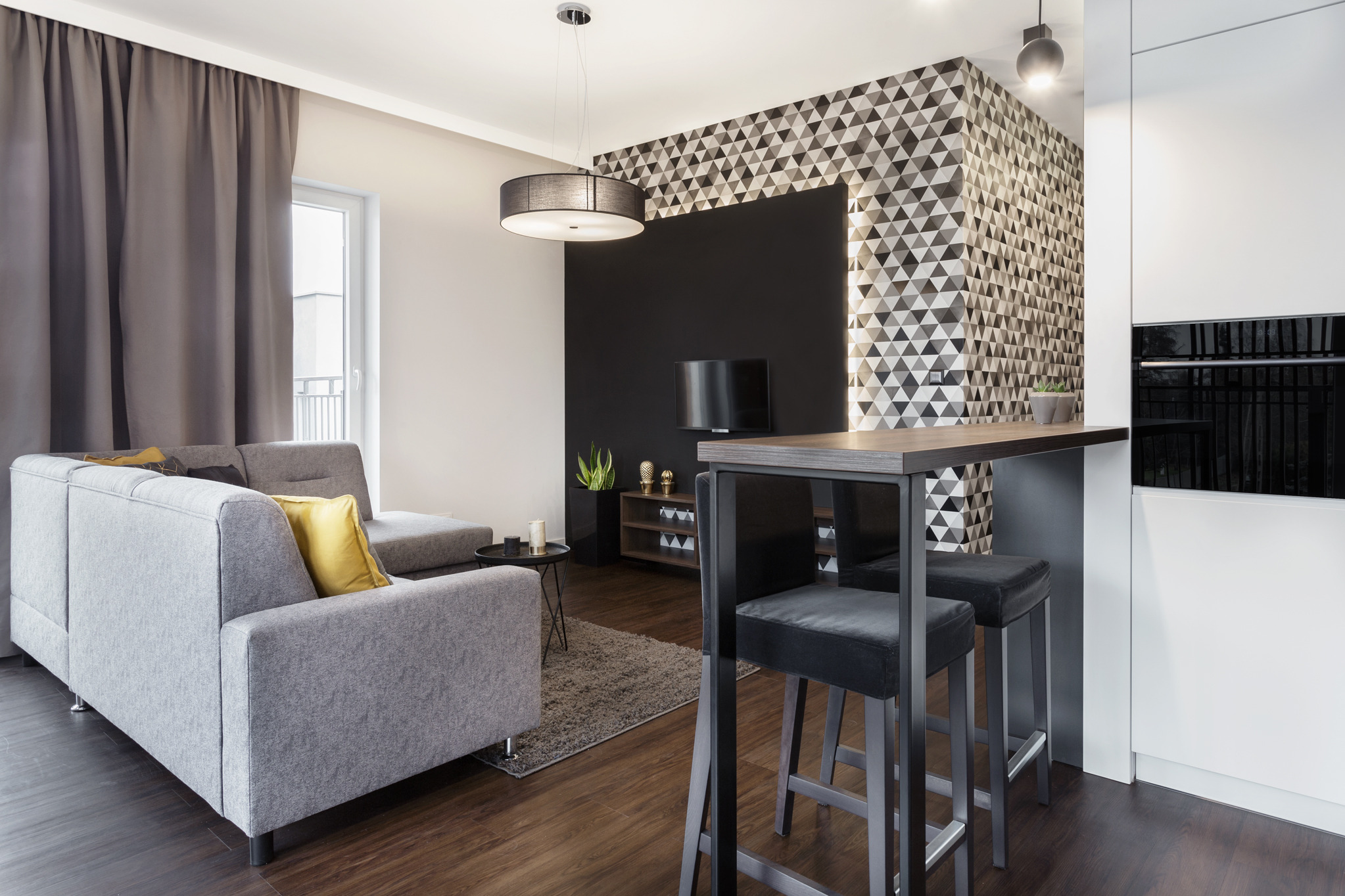 Nowoczesny salon z szarą sofą, geometryczną tapetą i drewnianym blatem baru z hokerami. Minimalistyczne oświetlenie i ciemna podłoga dodają elegancji.