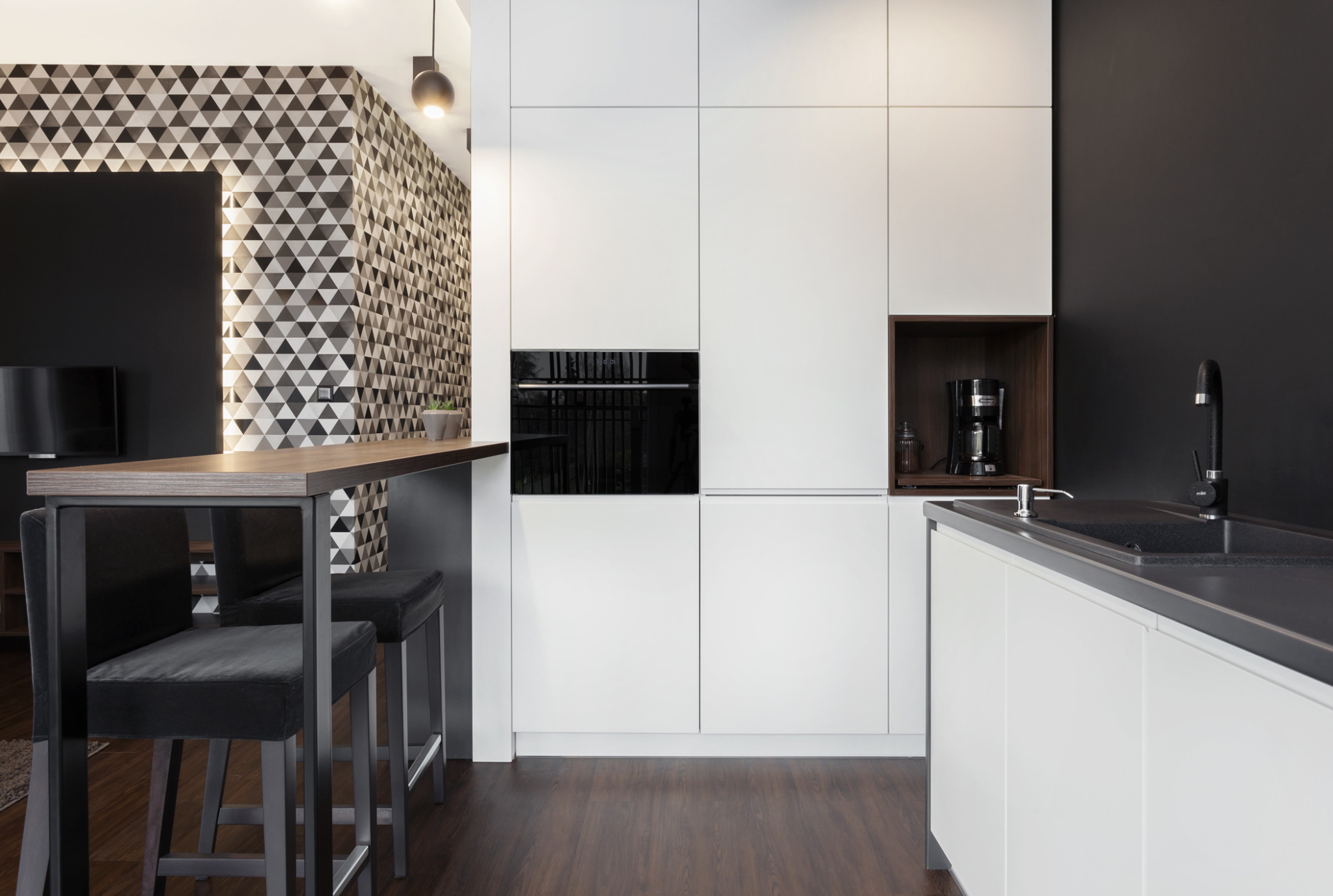 Nowoczesna kuchnia z białymi szafkami, czarnym blatem i geometryczną tapetą. Widoczne wysokie krzesła barowe i czarne elementy wyposażenia.