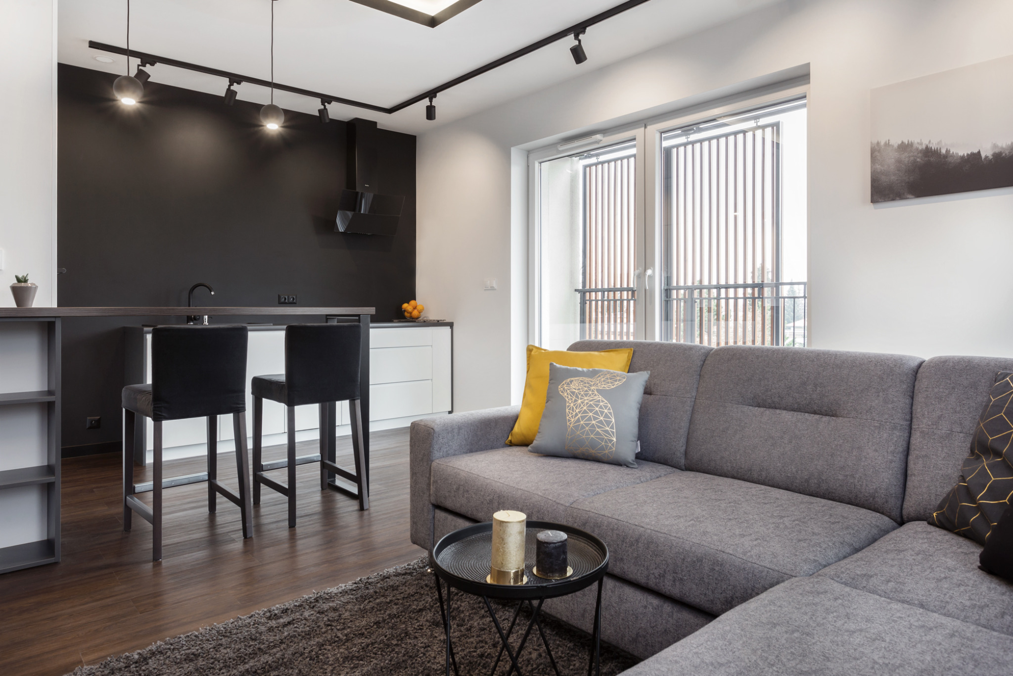 Nowoczesne wnętrze: szary narożnik z poduszkami, stolik kawowy z świecami, wyspa kuchenna z hokerami na tle czarnej ściany i białych szafek. Minimalistyczny design.