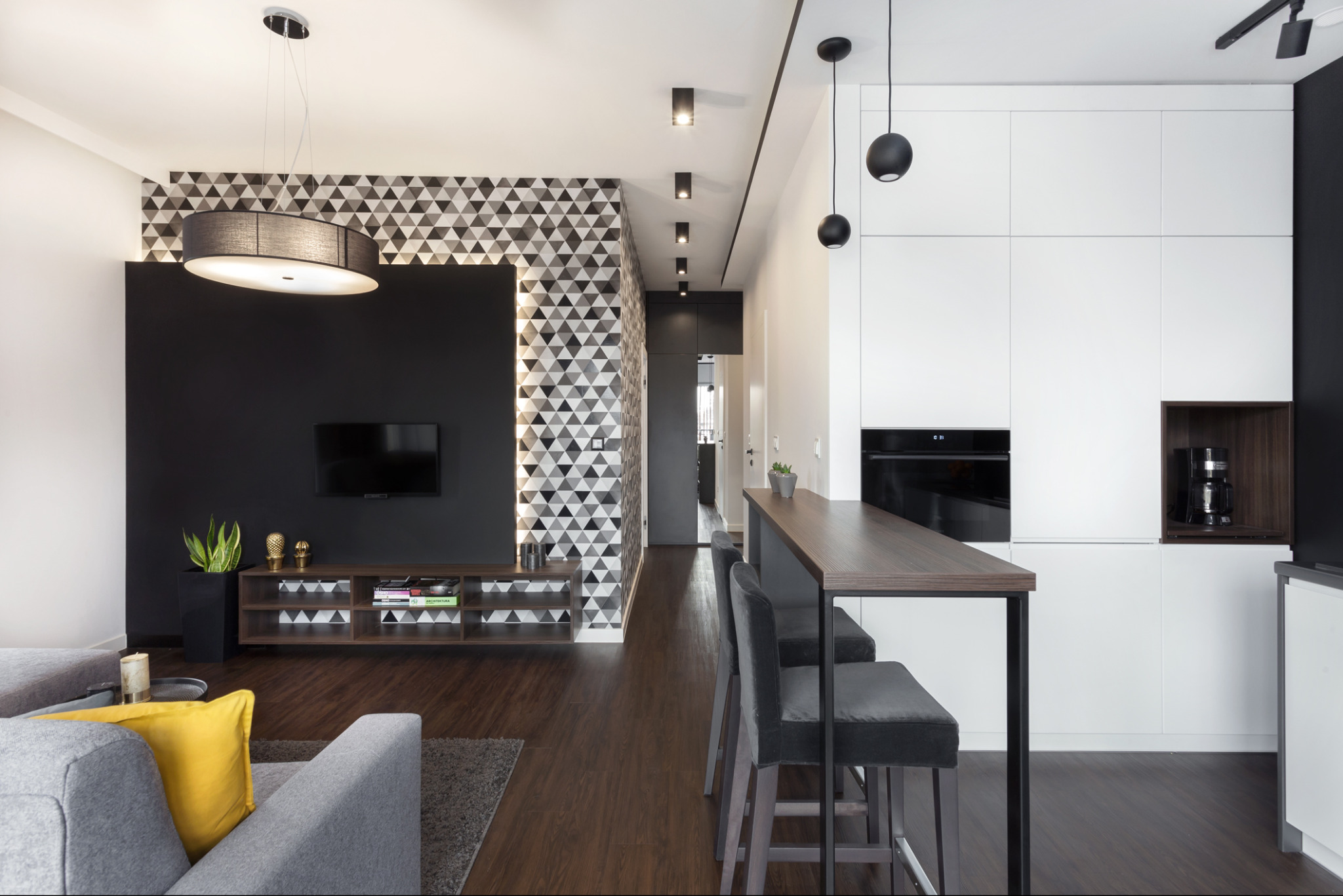 Nowoczesny salon z aneksem kuchennym w Siemianowicach Śląskich. Geometryczna tapeta, drewniana podłoga, szara sofa z żółtą poduszką i wysoki stół barowy.