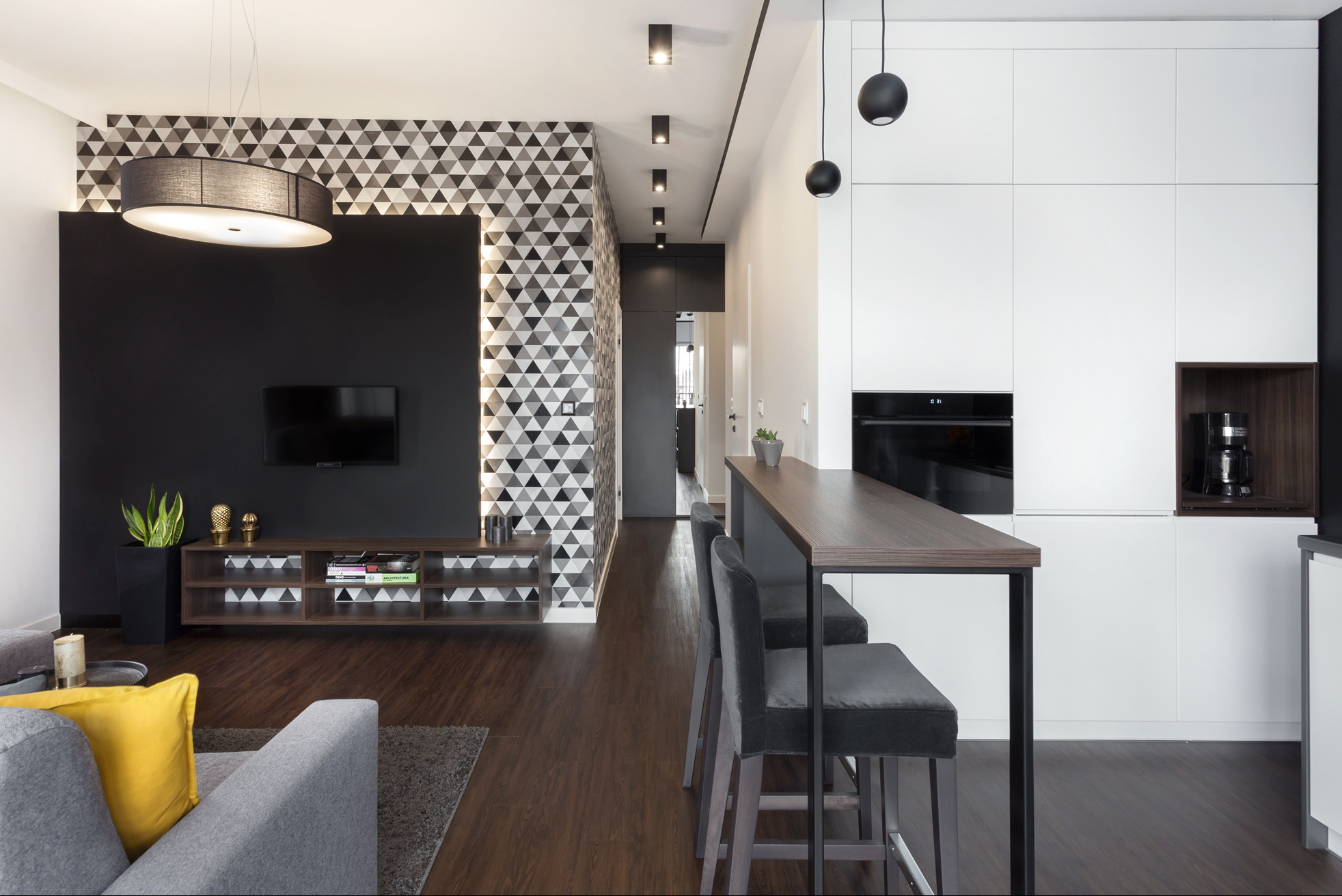 Nowoczesny salon z aneksem kuchennym: ciemna ściana z TV, geometryczna tapeta, drewniana podłoga i wyspa kuchenna z krzesłami barowymi.