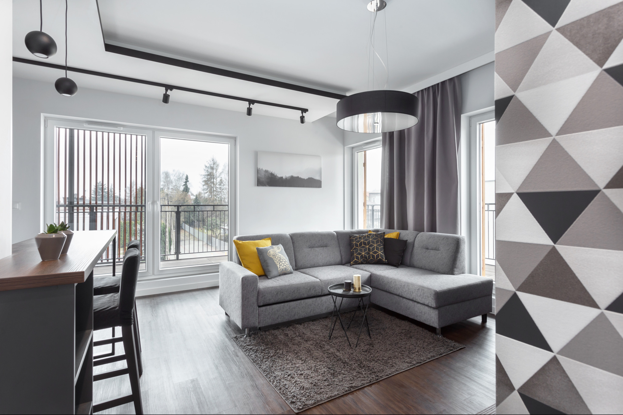 Nowoczesny salon z szarą sofą narożną, geometryczną tapetą i oknami balkonowymi. Minimalistyczne oświetlenie punktowe i wisząca lampa. Widok na balkon.
