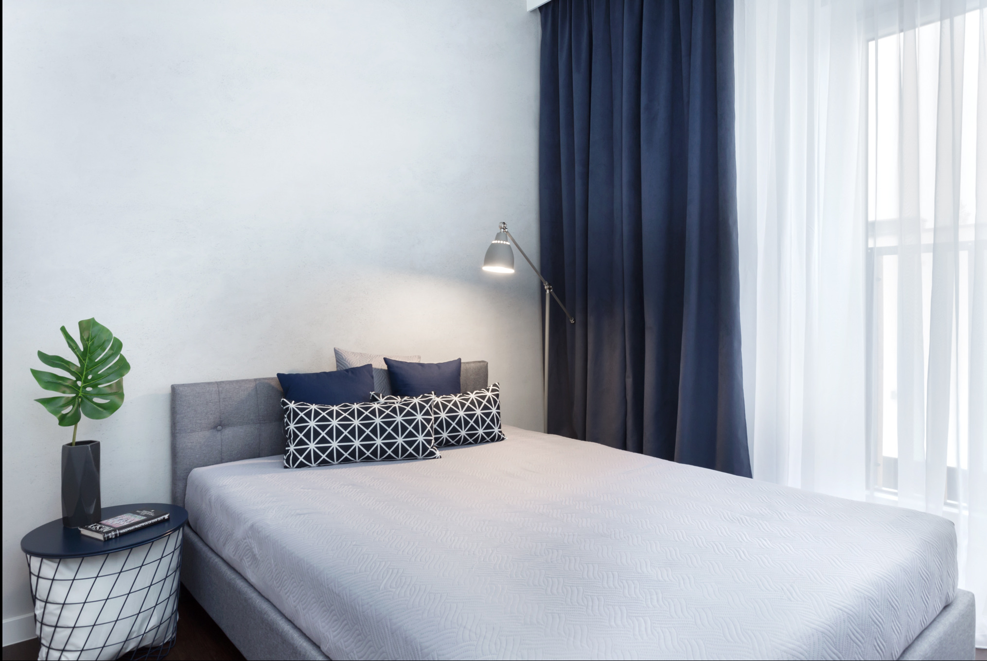 Jasna sypialnia z szarym łóżkiem, ciemnymi poduszkami i zasłonami. Na stoliku nocnym roślina w wazonie i książka. Lampa podłogowa doświetla wnętrze.