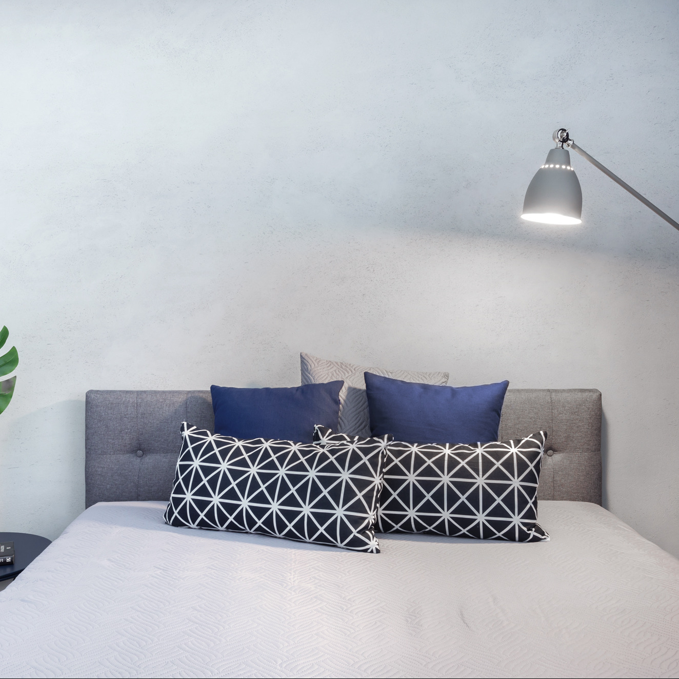 Minimalistyczna sypialnia z szarym zagłówkiem, niebieskimi i geometrycznymi poduszkami. Lampa podłogowa oświetla łóżko, a z boku widoczny jest stolik.