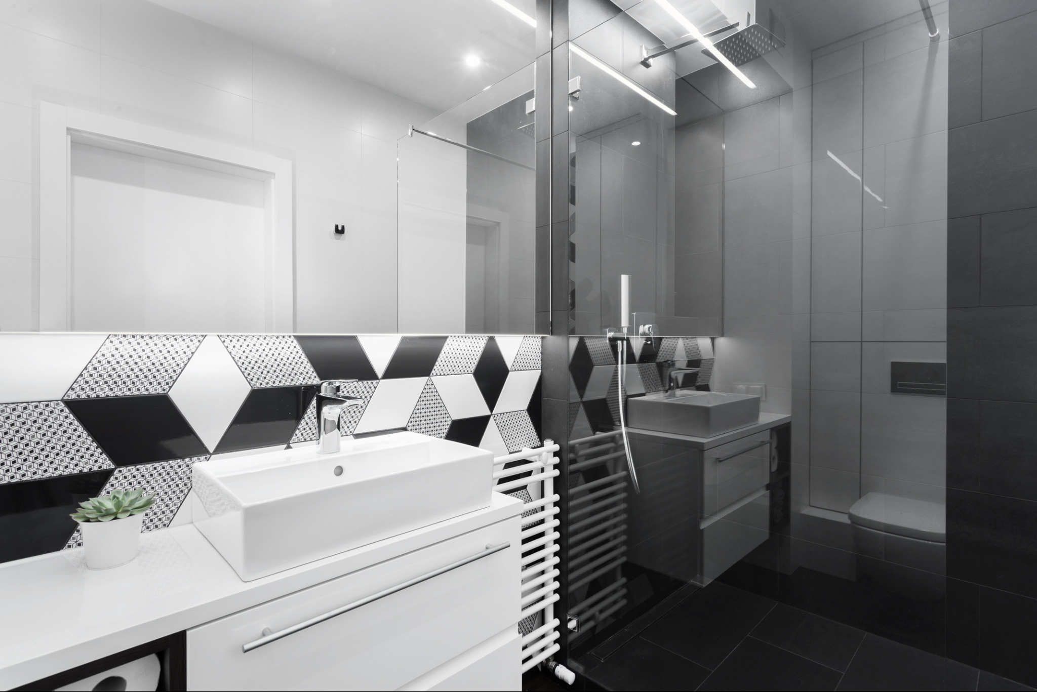 Nowoczesna łazienka z białą umywalką na szafce, wzorzyste płytki nad blatem, grzejnik drabinkowy i kabina prysznicowa ze szklaną ścianką.