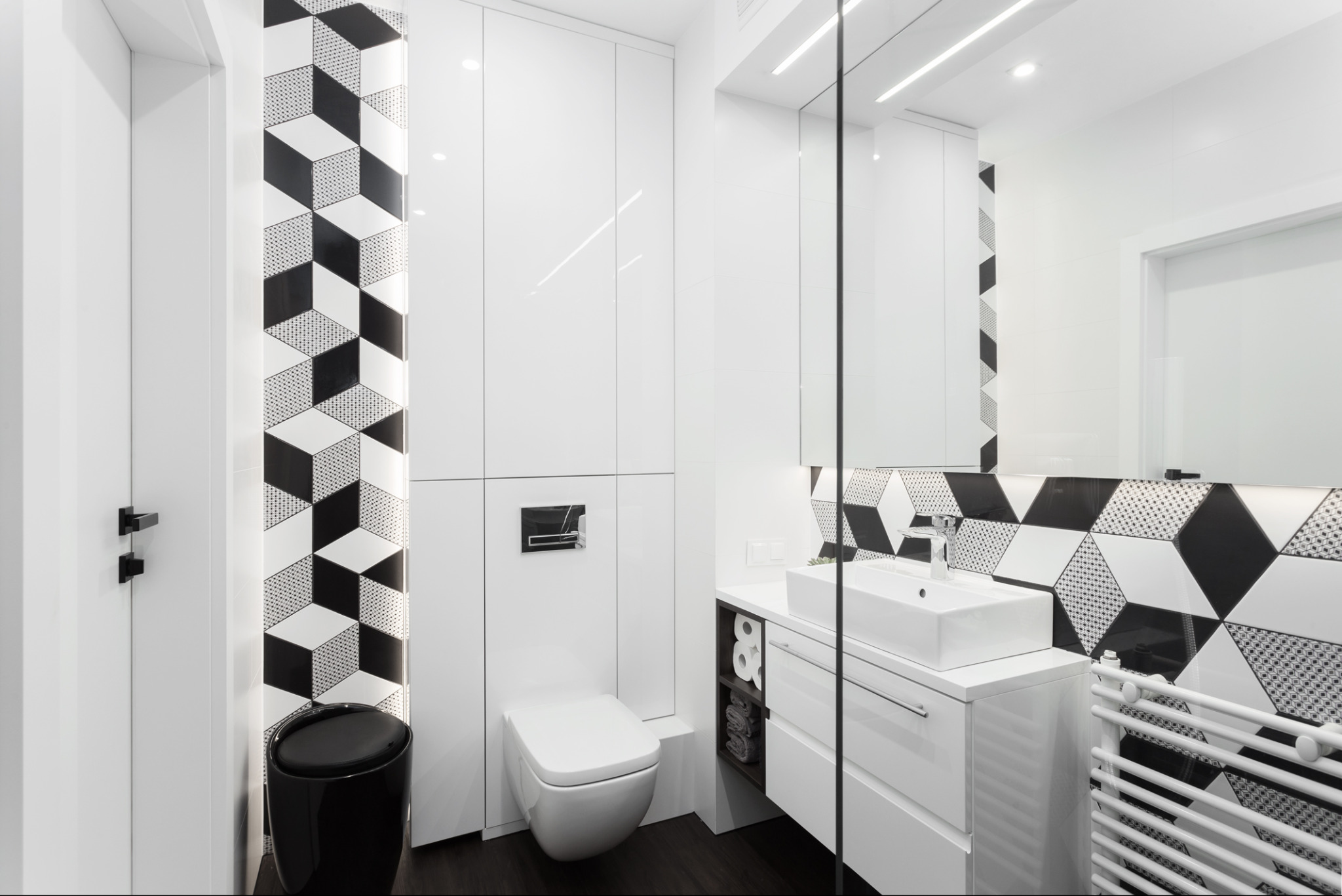 Nowoczesna, biała łazienka z czarnymi akcentami: geometryczne płytki, minimalistyczna umywalka, toaleta i grzejnik. Czyste linie, elegancki design, dobrze oświetlone wnętrze.