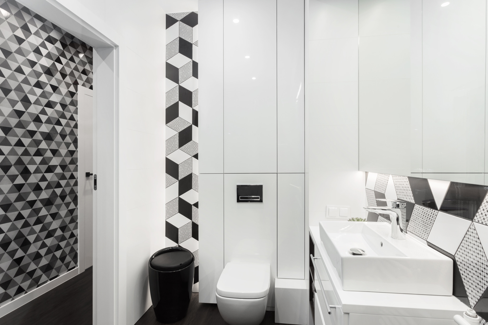 Nowoczesna łazienka z geometrycznym wzorem na ścianie, białą armaturą i szafkami w zabudowie. Czarny kosz na śmieci dodaje kontrastu.