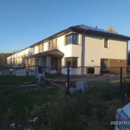 Nowe domy w zabudowie szeregowej z panelami słonecznymi na dachu, widoczne prace brukarskie przed posesją, ogrodzenie siatkowe.