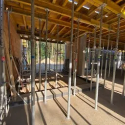 Wnętrze budynku w trakcie budowy z widocznymi podporami stropowymi, deskowaniem drewnianym oraz fragmentami ścian murowanych w tle.