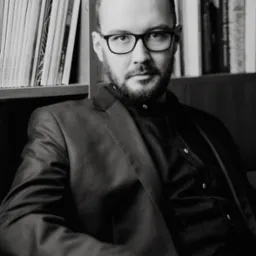 Portret mężczyzny w okularach i marynarce, siedzącego na tle regału z książkami, w czarno-białej tonacji.