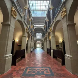 Długi korytarz w budynku sądu z rzędem kolumn, łuków i drewnianych ławek, wyłożony czerwoną heksagonalną terakotą z ciemniejszym geometrycznym wzorem na podłodze.