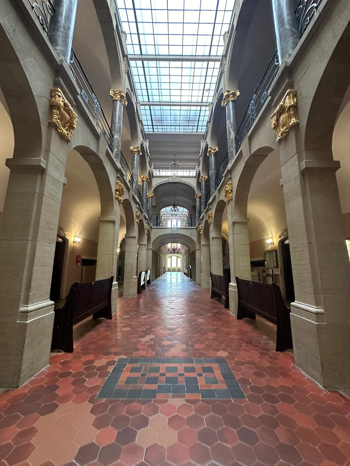 Długi korytarz w budynku sądu z rzędem kolumn, łuków i drewnianych ławek, wyłożony czerwoną heksagonalną terakotą z ciemniejszym geometrycznym wzorem na podłodze.
