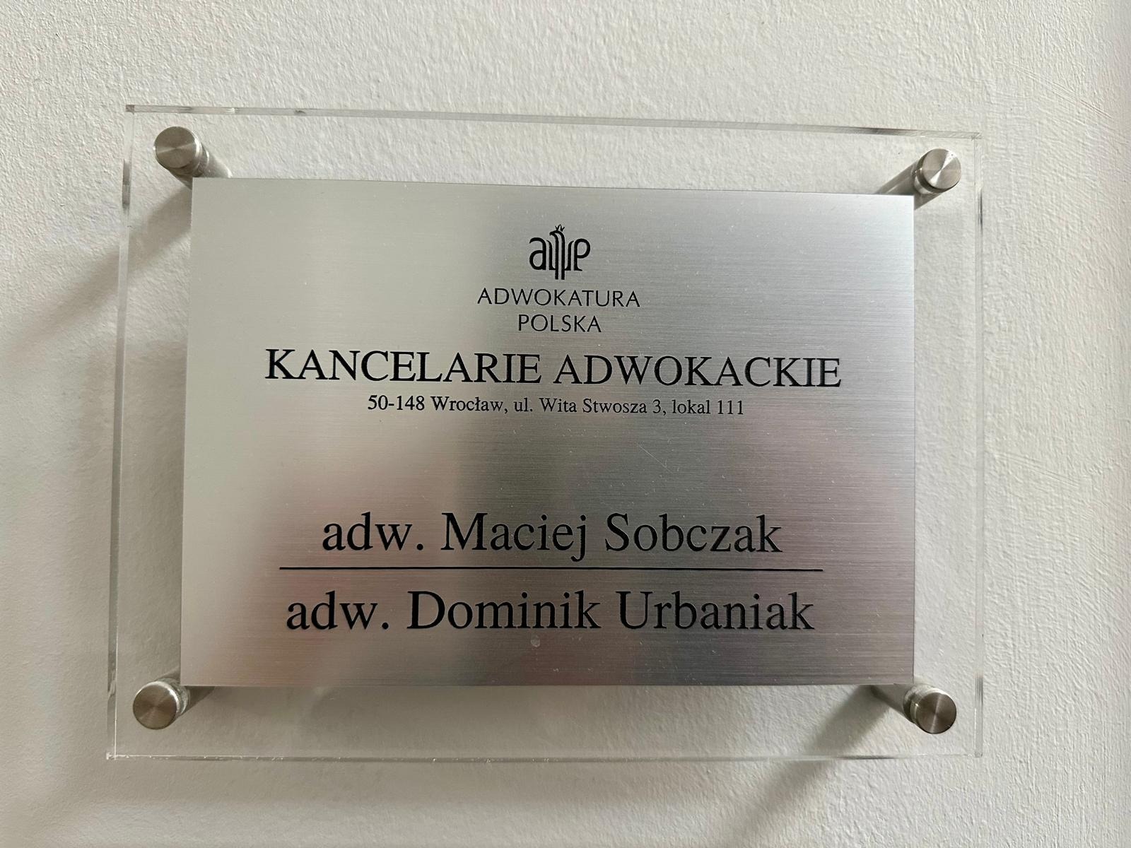 Tabliczka informacyjna z nazwą Kancelarii Adwokackiej, adresem we Wrocławiu i imionami adwokatów Macieja Sobczaka i Dominika Urbaniaka, zamontowana na ścianie.