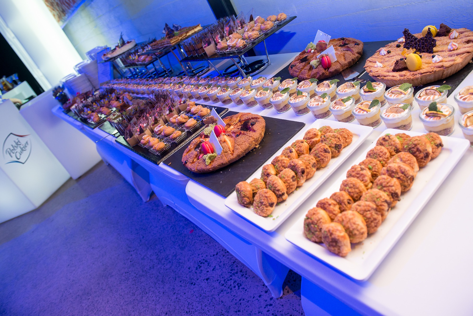 Stół z różnorodnymi przekąskami i deserami: tarty z owocami, mini desery w szklanych naczyniach, kruche ciasteczka i inne słodkości, przygotowane na przyjęcie.