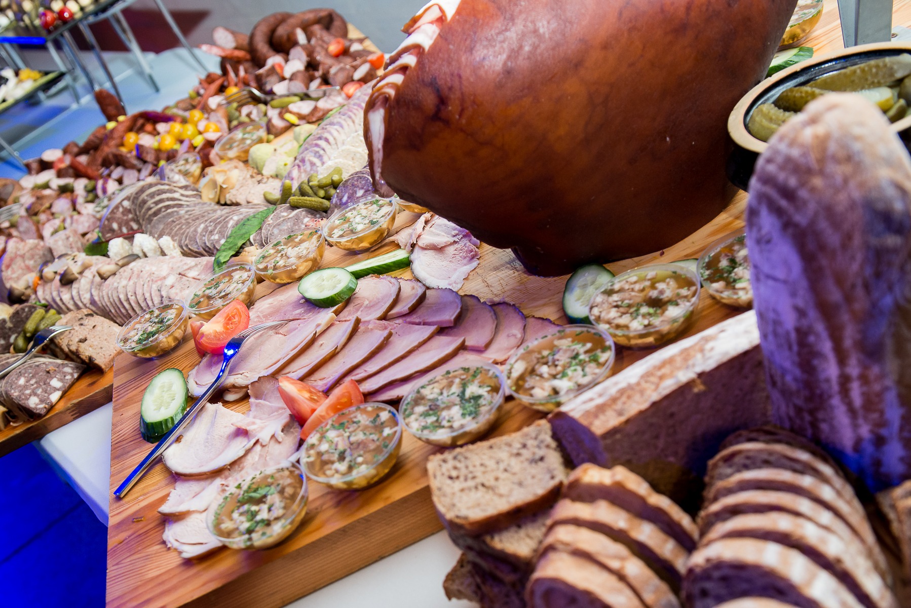 Bogato zastawiony stół z różnorodnymi wędlinami, pasztetami, pieczywem, ogórkami konserwowymi i galaretkami w małych miseczkach, tworzący apetyczny bufet.